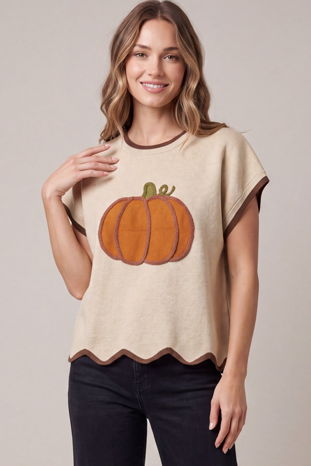 Model wearing a cream drop shoulder short sleeve sweater top with a pumpkin applique graphic on the front, scallop hem, and brown contrast trim at the neckline, sleeves, and hem, styled with dark jeans