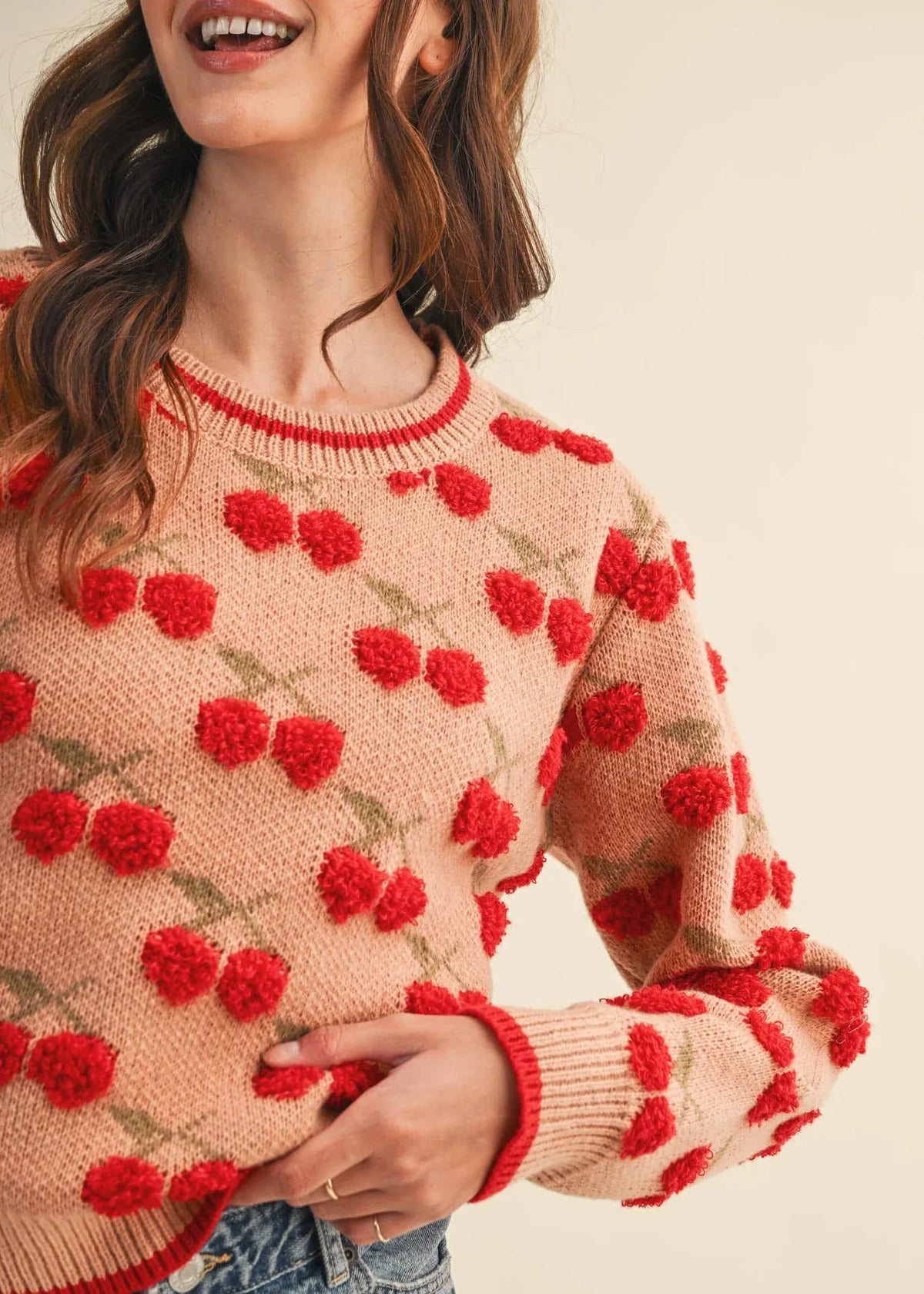 Model wearing a camel knit sweater with raised fuzzy red cherries, muted green stems, and red trim at the ribbed neckline, cuffs, and hem.