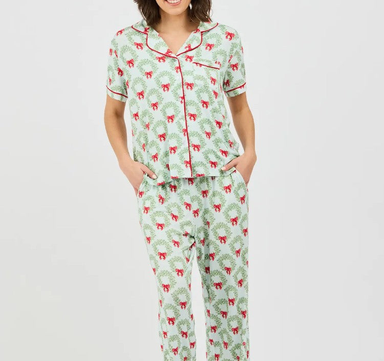 A woman modeling a matching short-sleeve pajama top and pants set with a festive holiday print of red Christmas trees on a light green background, featuring red piping details.