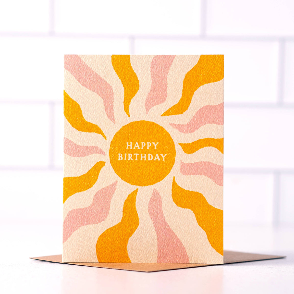 Boho sun birthday card with golden yellow and blush pink rays on a cream background, white “Happy Birthday” lettering, and a kraft envelope.
