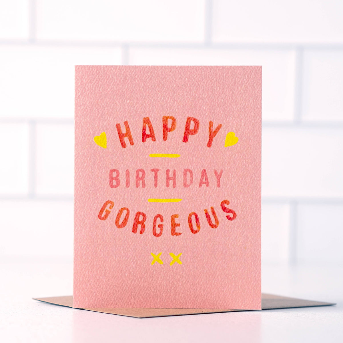 Pink “Happy Birthday Gorgeous” greeting card with red and coral lettering, yellow hearts and kiss marks, shown with a kraft envelope.