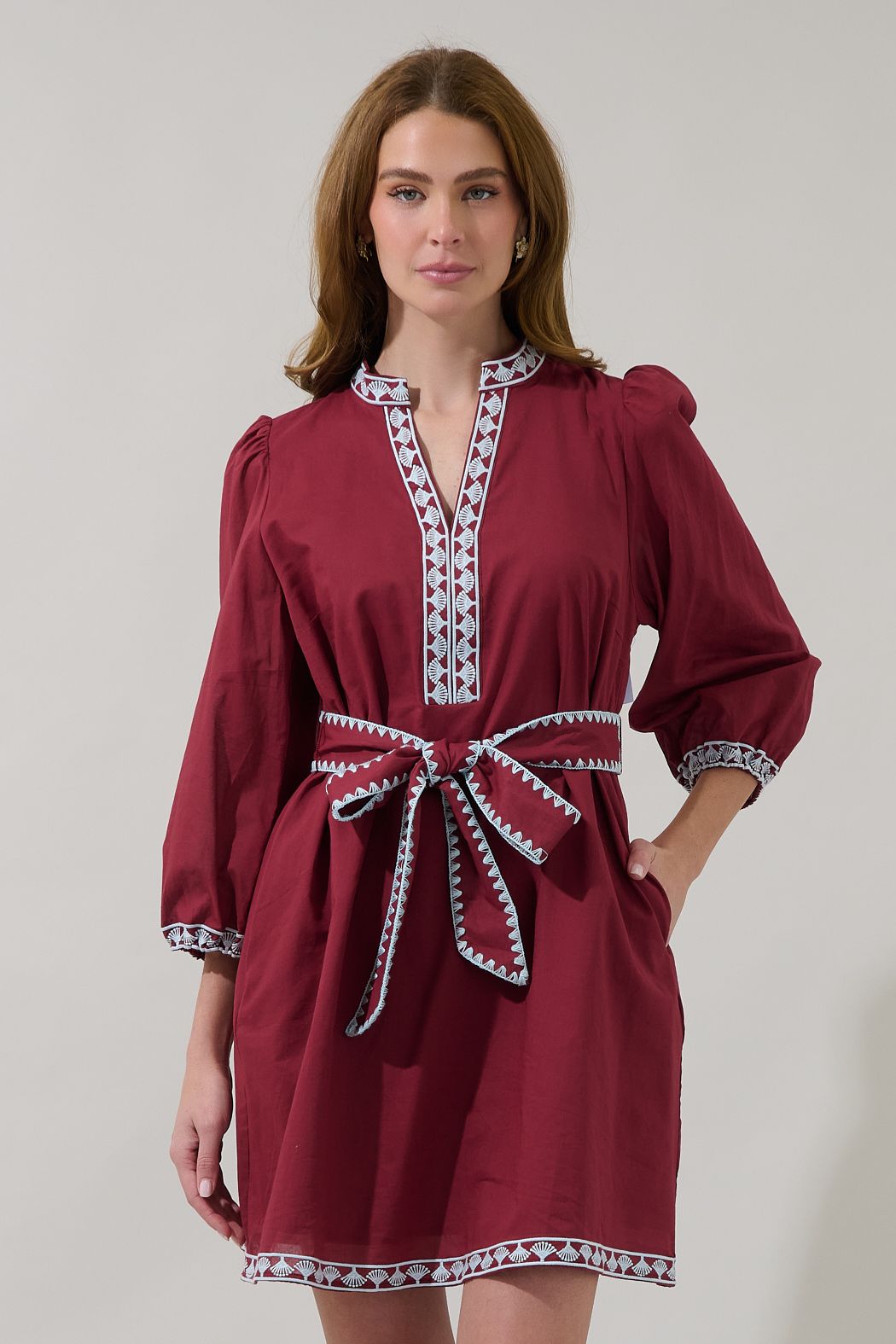 Model wearing a maroon shift dress with blue and white border embroidery, long sleeves, a V-neckline, and a matching tie belt