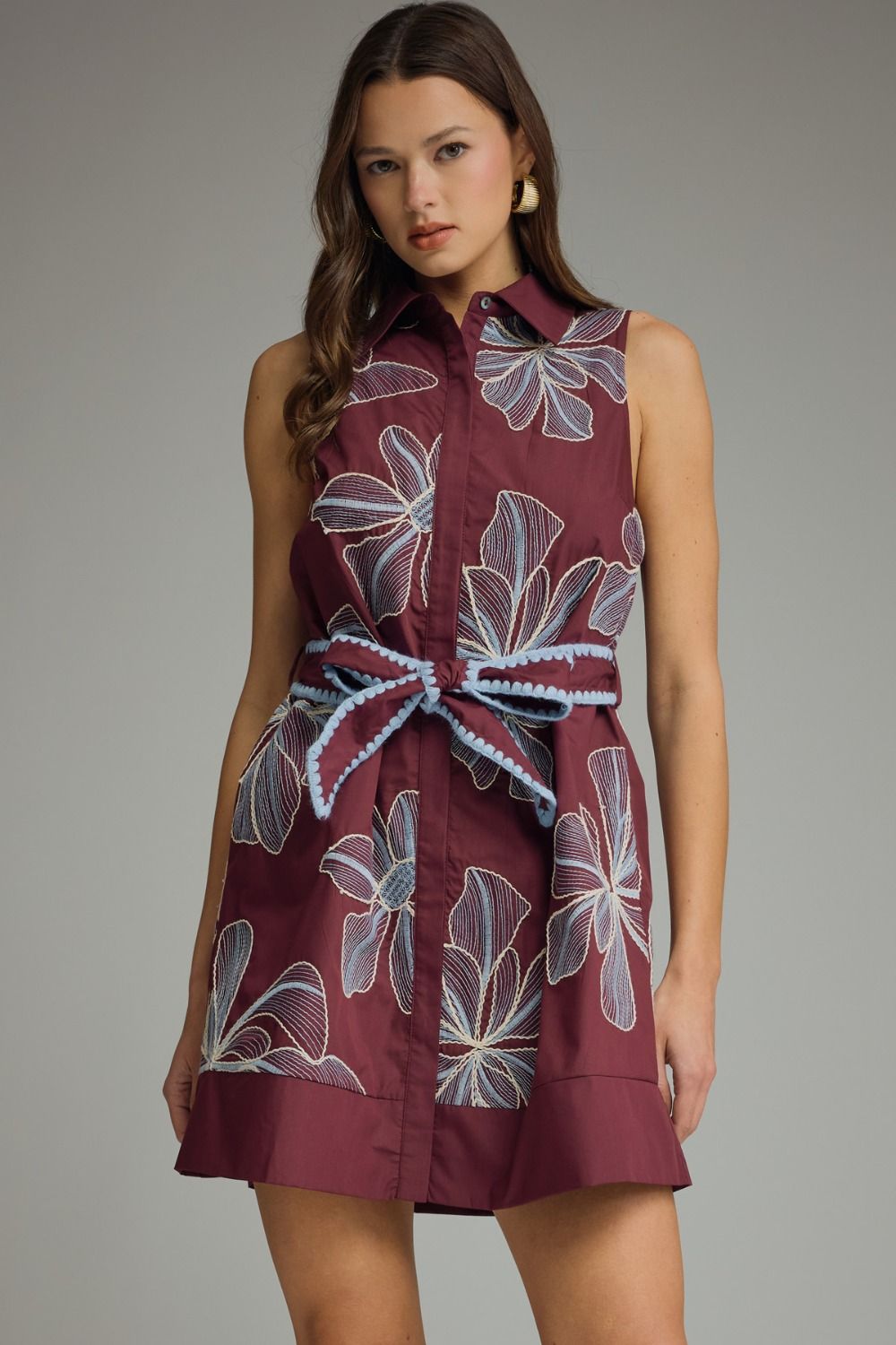Burgundy sleeveless mini dress with all-over white and blue sketch-style floral embroidery, collared neckline, button-front closure, self-tie waist belt with contrast pom-pom trim, A-line silhouette, side pockets, fully lined.