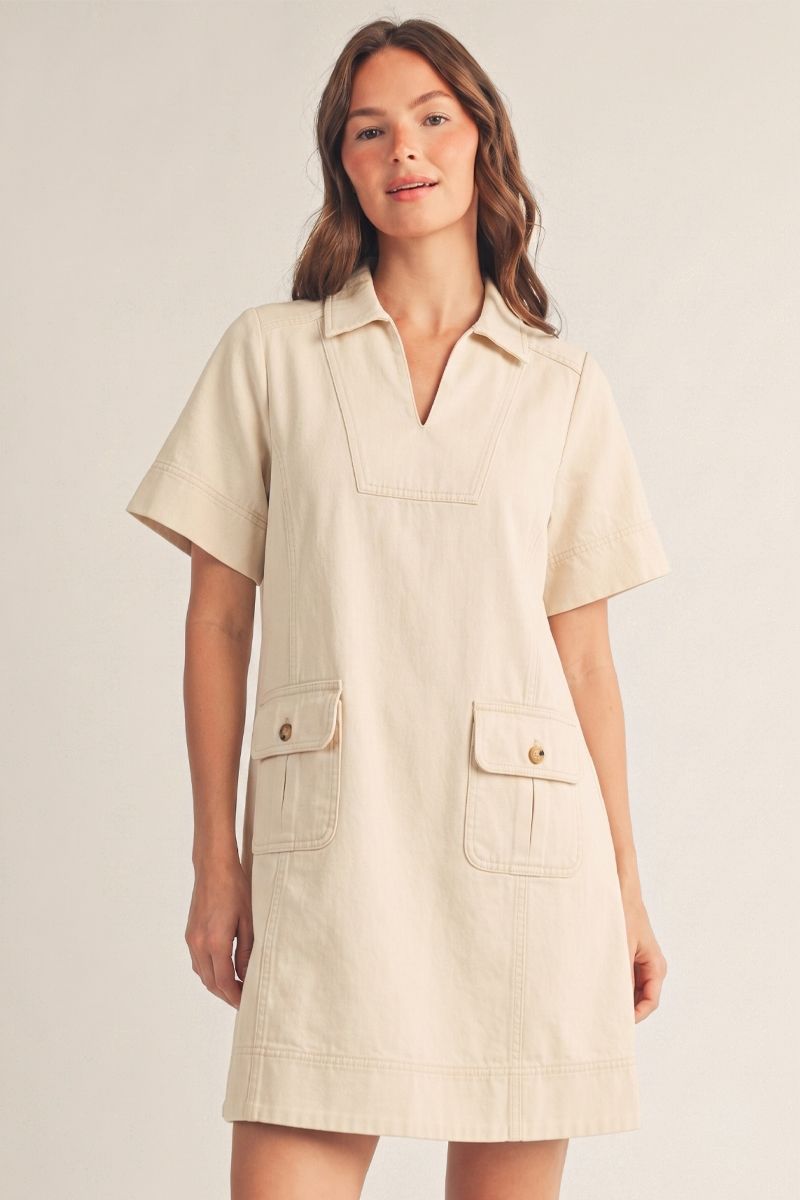 Oatmeal garment-washed shift dress with notched V-neckline, collared detail, short boxy sleeves, A-line silhouette, functional side pockets with button closures, chest pocket, visible topstitching, and horizontal seam detail near hem.