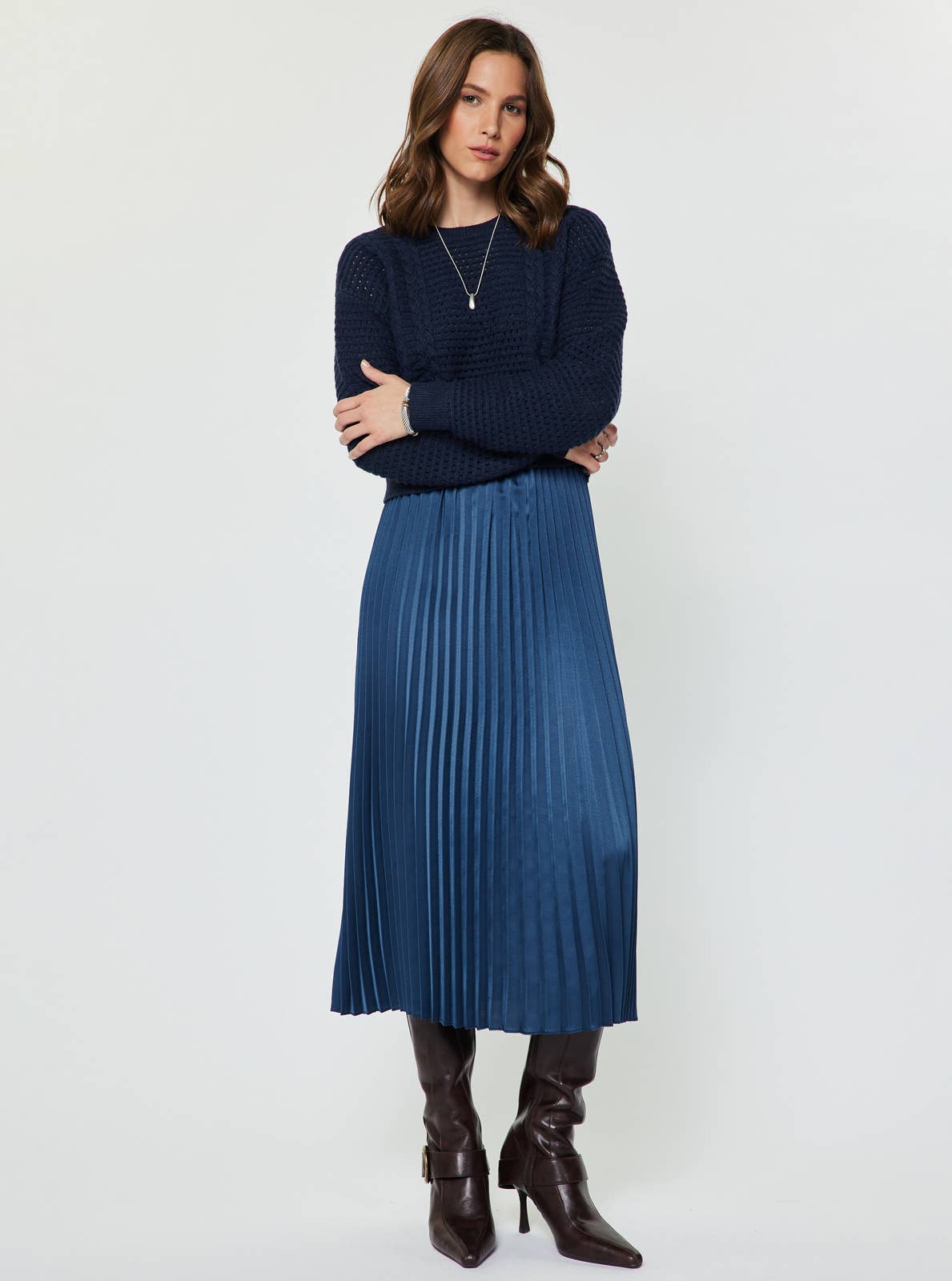 Model wearing a navy cable knit pullover over a blue satin pleated midi dress, styled with dark knee-high boots.