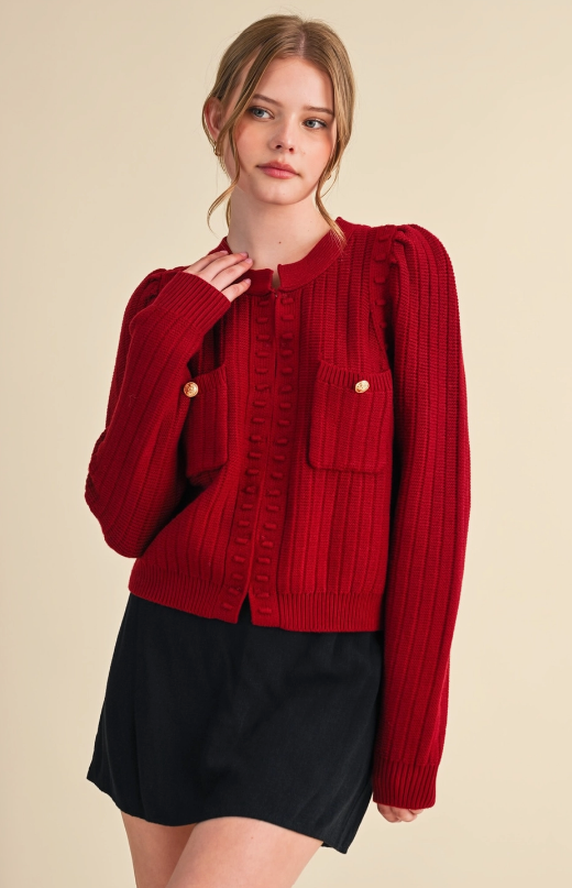 Model wearing a burgundy textured knit cardigan with puff shoulders, front patch pockets, and gold-tone pocket buttons.