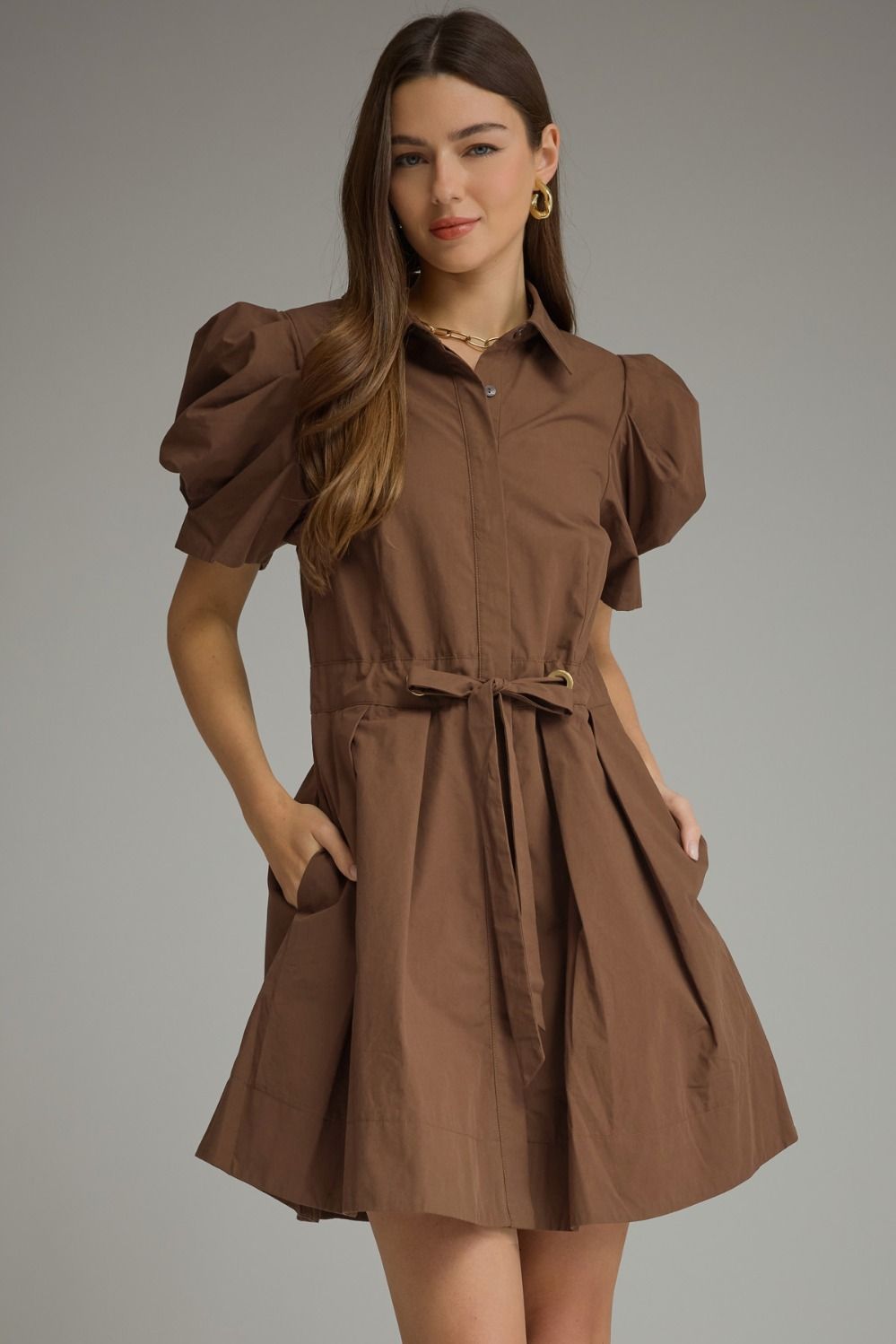 Model wearing a chocolate brown mini shirt dress with short puff sleeves, a collared neckline, tie waist, pleated skirt, and side pockets.