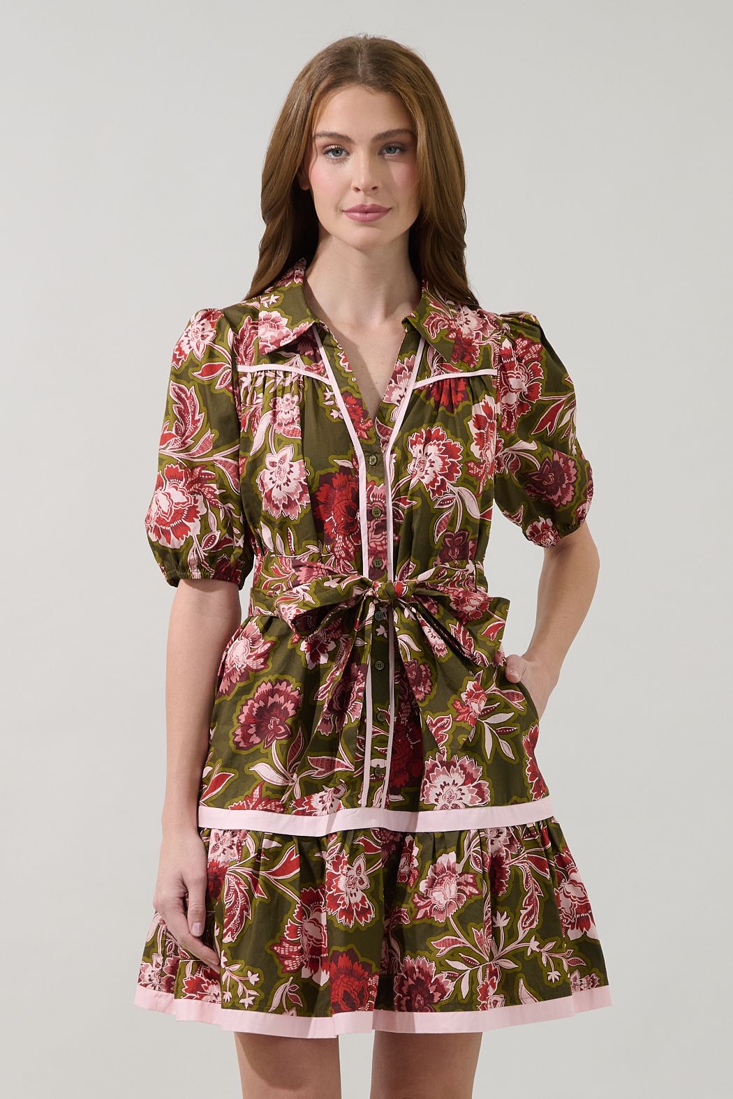 Model wearing a green and pink floral shift mini dress with a collared button front, puff sleeves, contrast trim, waist tie, and tiered skirt