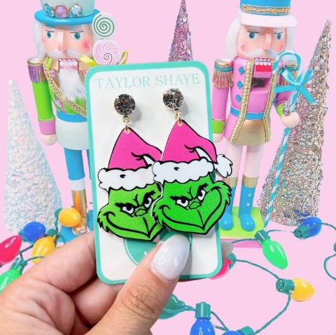 Grinch face dangle earrings with green faces, pink Santa hats, white hat trim, and textured round metallic tops, displayed on a jewelry card.