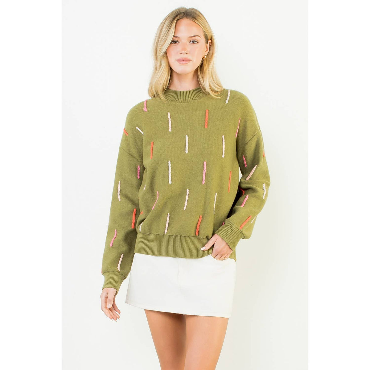 Model wearing an olive green knit sweater with pink, orange, and cream dash details, dropped shoulders, and ribbed neckline, cuffs, and hem.⠋
