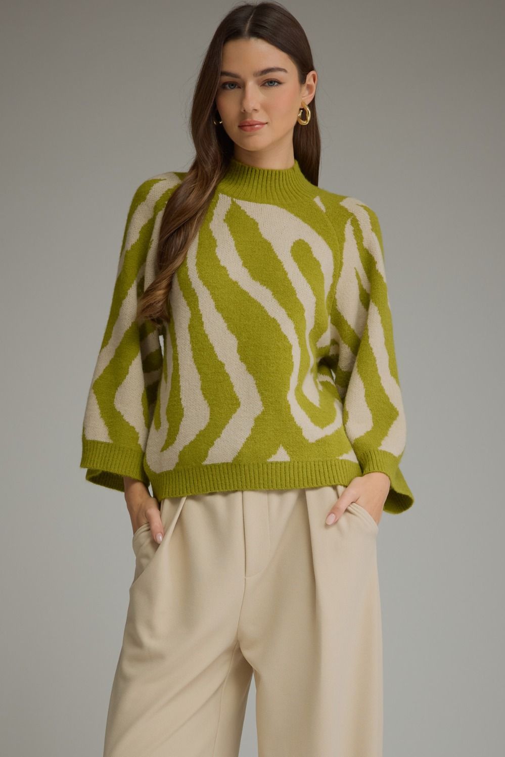 Model wearing an olive green and cream abstract print mock neck sweater with three-quarter bell sleeves and cream trousers.