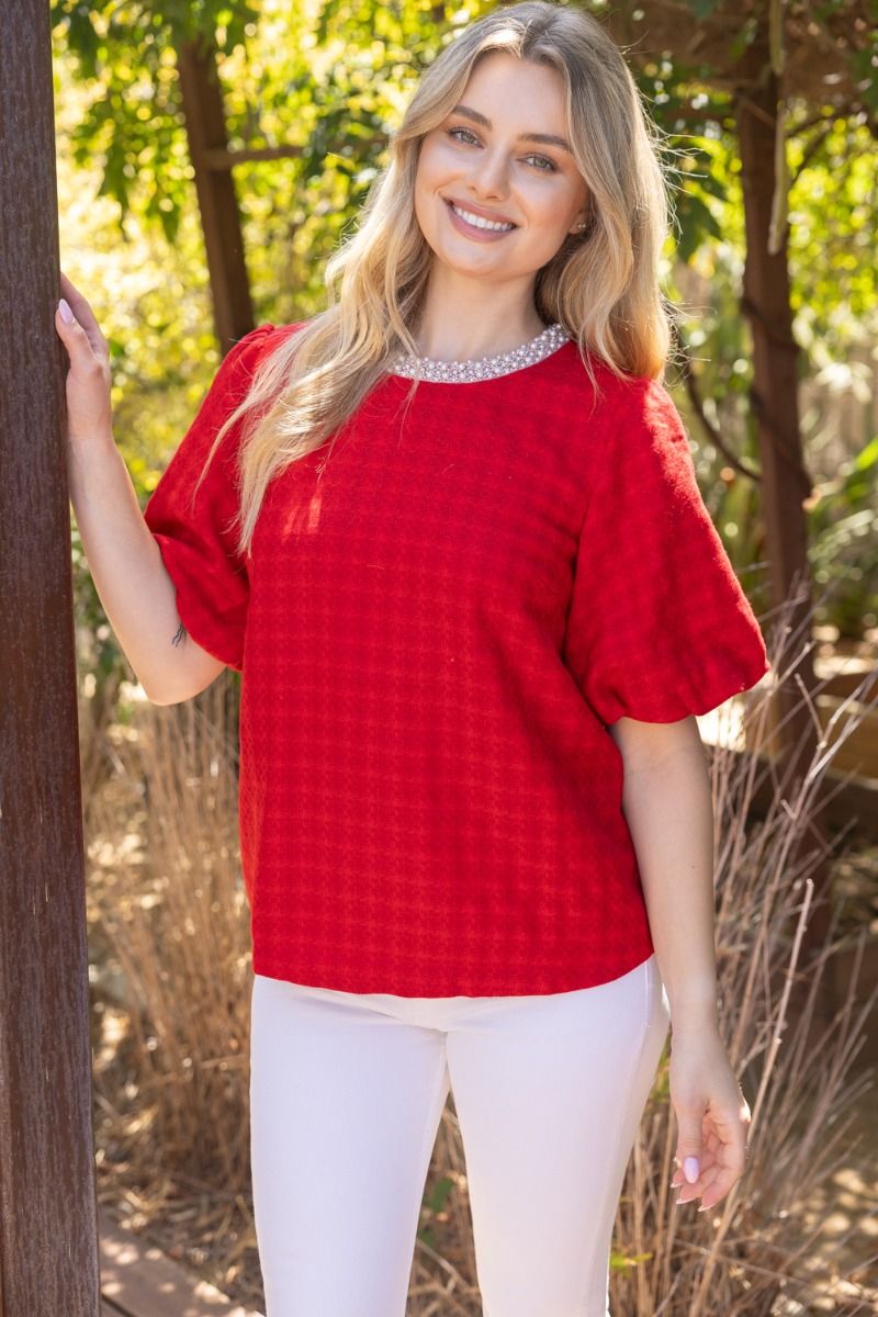 Red houndstooth bubble sleeve top with pearl neckline detail on model styled with white trousers
