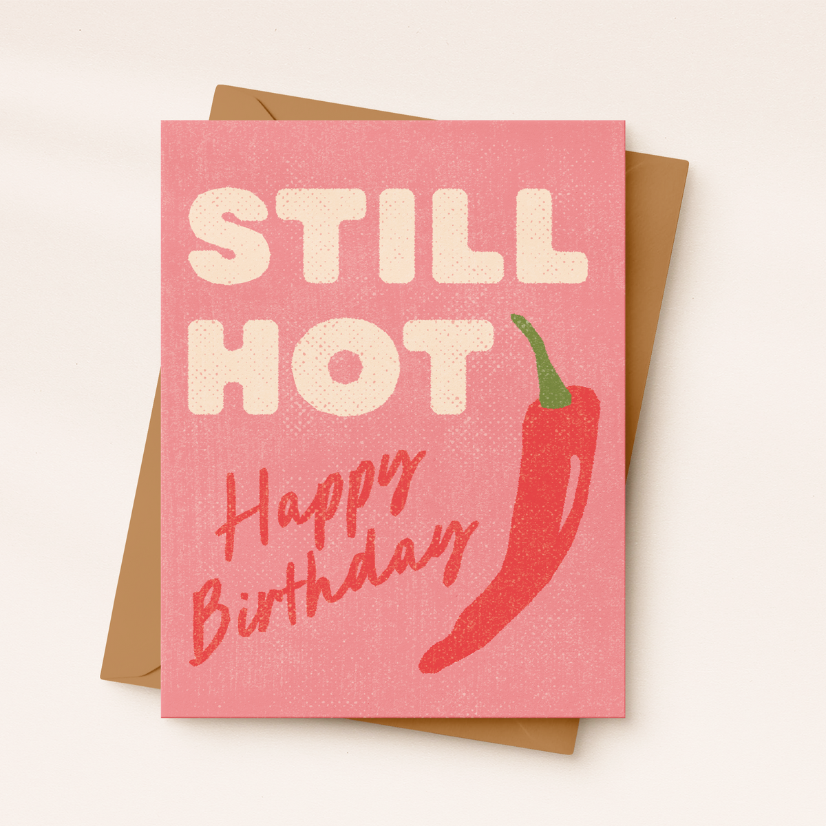 Pink birthday card with cream “Still Hot” lettering, a red chili pepper, and red “Happy Birthday” script, shown with a kraft envelope.