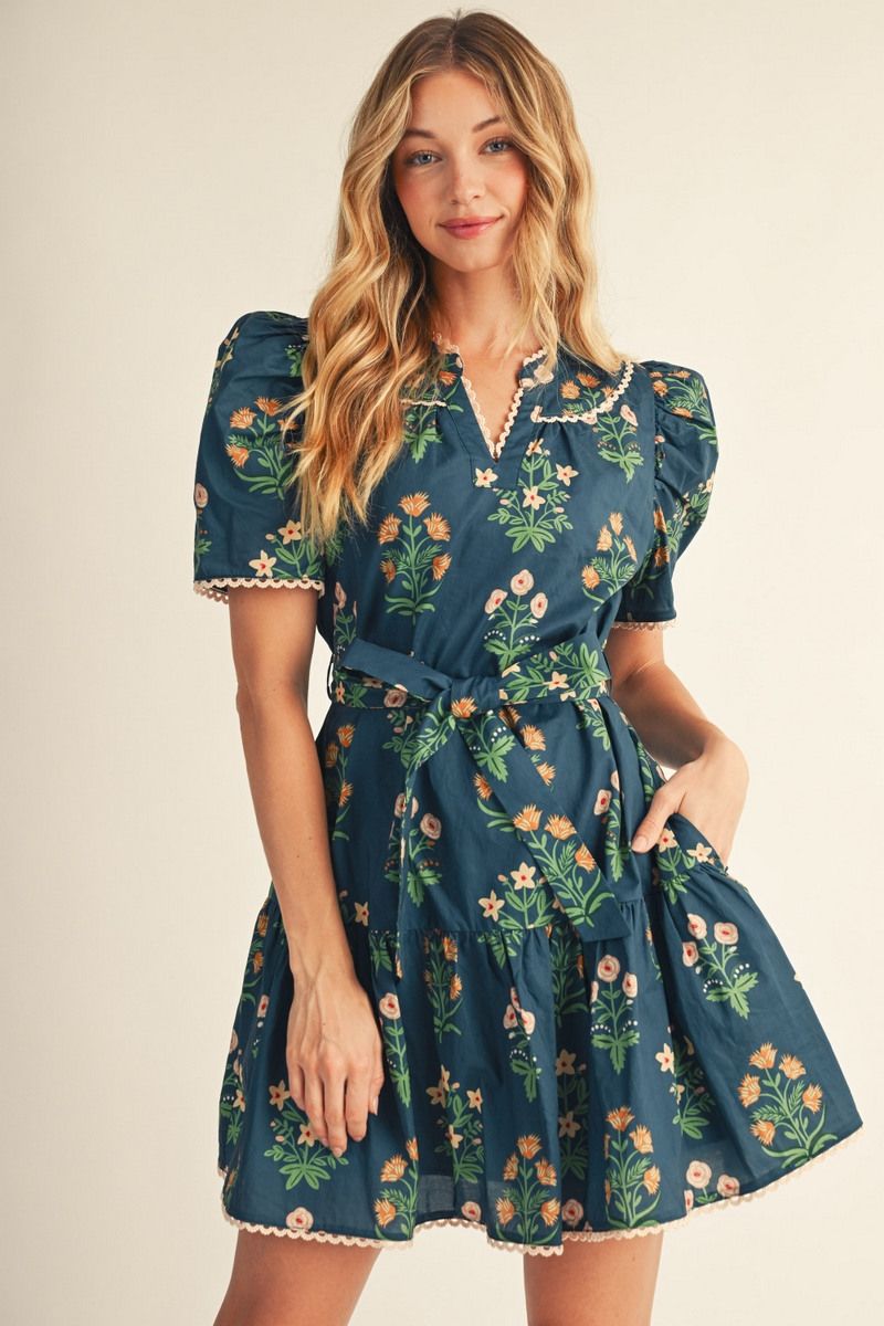 Dark teal floral dress with multicolor botanical print, V-neckline with scalloped lace trim, puffed short sleeves with scallop trim at cuffs, fitted waist with tie-belt closure, A-line skirt with side pockets, and scalloped lace trim along hemline.
